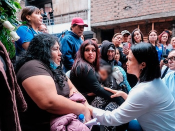 Grupo de personas en una calle estrecha. Una mujer de camisa blanca interactúa con otra mujer. Se ven rosas blancas y edificios de ladrillo al fondo