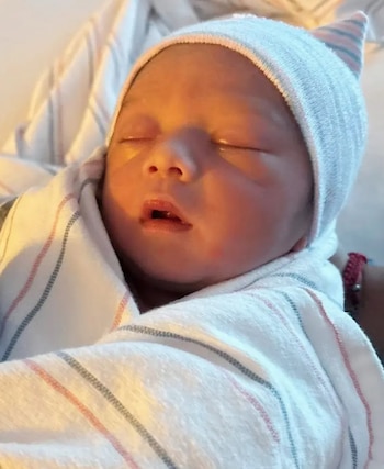 Primer plano de un bebé recién nacido dormido, envuelto en una manta blanca con rayas finas rosadas y azules, usando un gorro a juego