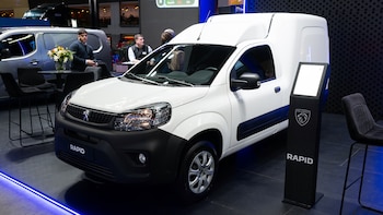 Una van Peugeot Rapid blanca en exposición. Stand negro con logo, dos hombres, sillas, mesa con flores y una van gris en segundo plano