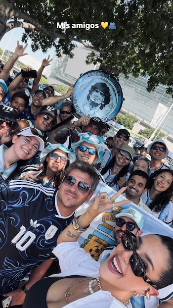 Un grupo numeroso de personas sonrientes, con camisetas, sombreros y una bandera de Argentina. Se ve un bombo con la cara de Maradona y árboles