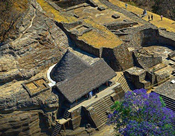 Fotografía aérea de ruinas arqueológicas con muros de piedra y estructuras de techos de paja. Muestra escaleras, caminos, vegetación y personas