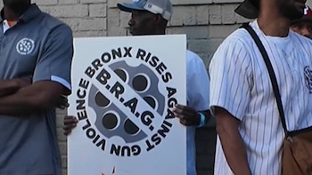 Primer plano de un hombre sosteniendo un cartel blanco con letras negras que dicen "BRONX RISES AGAINST GUN VIOLENCE B.R.A.G." y un tambor de revólver tachado