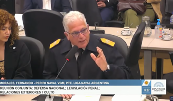 Un hombre con uniforme y gafas habla ante un micrófono en un salón de reuniones con un zócalo informativo en la pantalla