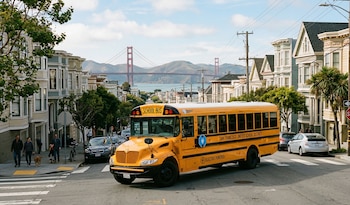 Autobús escolar amarillo eléctrico de San Francisco en una calle urbana con casas, árboles, personas y el puente Golden Gate a lo lejos
