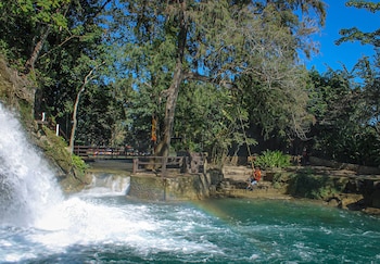 Una cascada vierte en un río turquesa con un arcoíris. Un hombre con chaleco salvavidas se balancea en una cuerda cerca de árboles y una cerca de madera