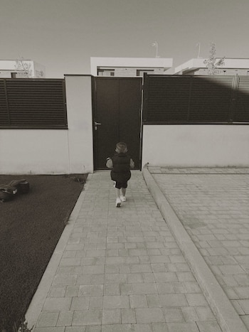 Fotografía monocromática de un niño de espaldas caminando sobre un sendero de baldosas. Al lado hay césped artificial y en el fondo se ven edificios blancos