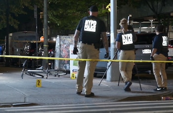 Tres agentes de la Policía de Nueva York detrás de una cinta policial amarilla con una bicicleta caída y marcadores de evidencia en una calle de noche