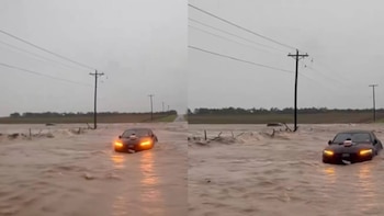 Coche oscuro con luces encendidas en carretera inundada con agua marrón. Postes de electricidad con cables y cielo nublado