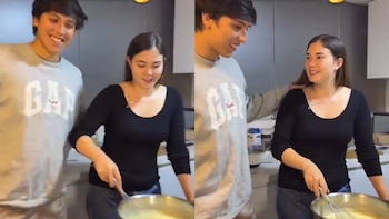 Kyara Villanella, con blusa negra, revuelve una olla en la cocina junto a Adrián Harboe, quien sonríe. Ambos jóvenes adultos