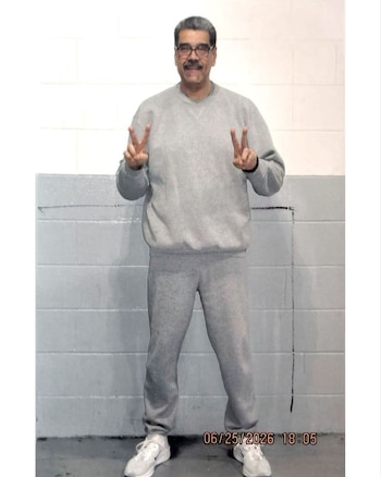 Hombre con bigote y anteojos viste ropa deportiva gris y zapatillas blancas mientras hace el gesto de la victoria con ambas manos