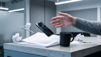 Una mano de adulto arroja un teléfono móvil sobre un escritorio de hormigón. Sobre el escritorio hay una pila de papeles, una taza, un bolígrafo y dos papeles arrugados.