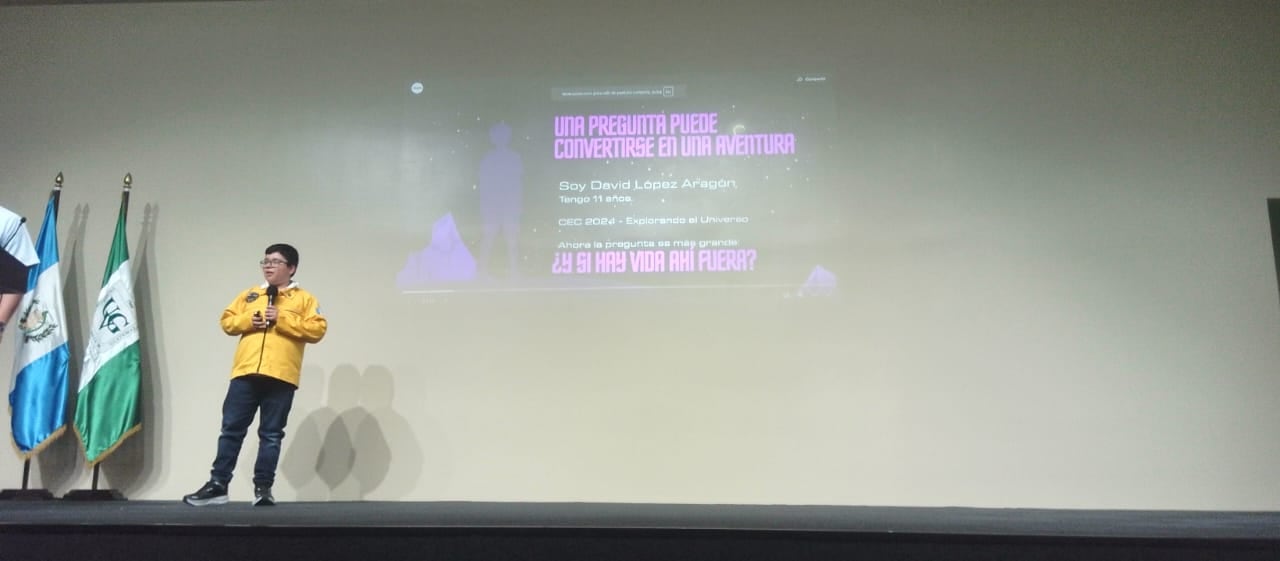 David López Aragón, de 11 años, presenta su ponencia sobre la exploración del universo junto a las banderas institucionales en un escenario. (Cortesía: Pamela López)