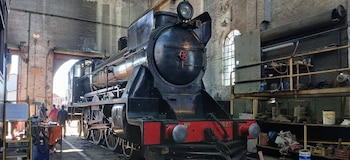 Una locomotora de vapor negra estacionada dentro de un taller industrial junto a herramientas, repuestos y tres personas