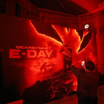 Una persona toma una foto con un teléfono a un hombre que posa sosteniendo un arma ficticia ante un panel rojo con el texto Gears of War E-Day