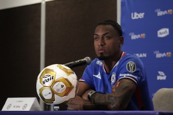 Hombre con camiseta azul de Cruz Azul y brazos tatuados cruza los brazos, mirando hacia adelante en una conferencia de prensa con un balón de fútbol