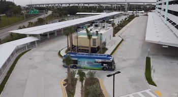 Vista aérea de un centro de transporte moderno con un autobús azul y verde, cubiertas blancas, palmeras y una autopista elevada en el fondo