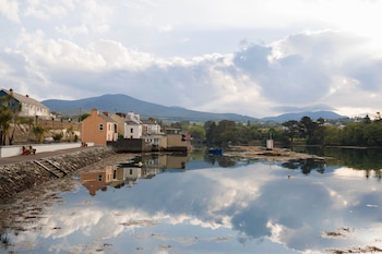 Pueblo costero con casas en la orilla de un cuerpo de agua. El agua refleja nubes y montañas. Se ve un bote azul y un poste blanco y rojo en el agua