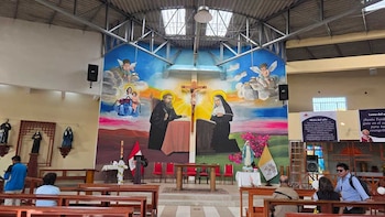 Bancas de madera frente a un altar con sillas rojas, una cruz, banderas, un mural religioso en la pared y techo de vigas metálicas