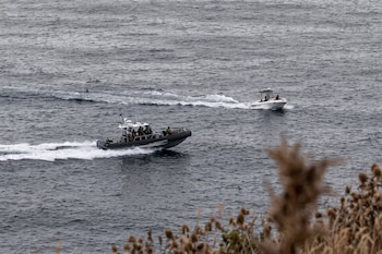 Dos embarcaciones, una de la Guardia Civil con tripulantes, y una lancha blanca, navegan en el mar. Matorrales secos en primer plano desenfocado