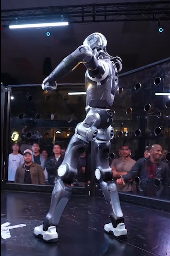 Robot humanoide de color gris metálico en postura erguida sobre una plataforma negra con espectadores al fondo