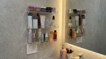 Estantes de acrílico llenos de productos de cuidado personal en una pared gris. Un espejo, tomacorriente, dispensador de jabón y cepillos de maquillaje son visibles.