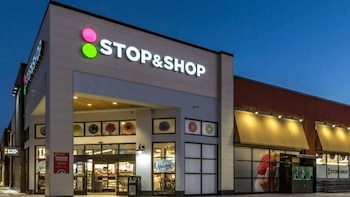 O exterior da loja Stop & Shop ao entardecer, com letreiros luminosos, toldos amarelos, grandes montras e a entrada principal visível.