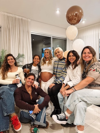 Ocho mujeres sonriendo en un salón moderno. Una mujer embarazada muestra su vientre. Hay globos marrones, beige y blancos al fondo