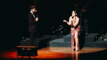 Adrián Bello en traje de vestir sostiene un micrófono mientras Daniela Darcourt, con vestido estampado, canta en un escenario iluminado