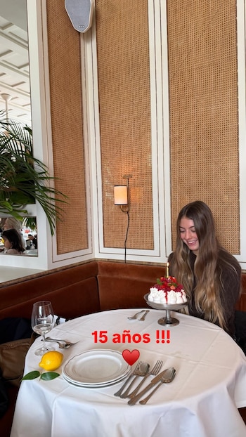 Una joven sonríe frente a un pastel con frutos rojos y una vela encendida en una mesa con cubiertos, vajilla y un limón