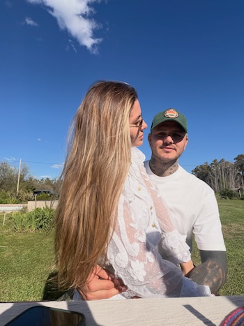 Un hombre con gorra verde y una mujer con blusa blanca de encaje posan al aire libre en un jardín