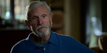 Un hombre de edad avanzada con cabello y barba grises usa un polo azul y una camiseta gris. El fondo está desenfocado y oscuro