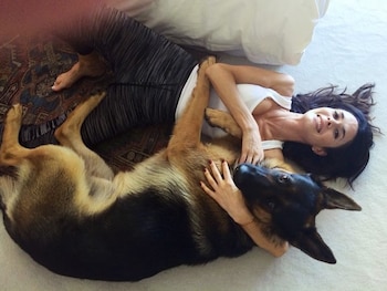 Rossana Almeyda, acostada en el suelo, sonriendo, con cabello oscuro suelto. Un perro pastor alemán recostado a su lado, ella lo acaricia con una mano