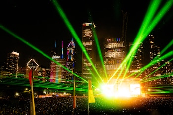 Multitud en un concierto, escenario con pantallas iluminadas, múltiples haces de luz láser verde, rascacielos, edificios con luces, cielo nocturno, banderas