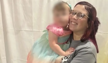 Una mujer con gafas y cabello oscuro rojizo sonríe mientras sostiene a un niño pequeño con un vestido rosa y azul. El rostro del niño está borroso