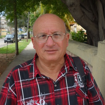 Primer plano de un hombre calvo de mediana edad con gafas y una camisa de cuadros roja y negra, con un sendero y árboles verdes de fondo