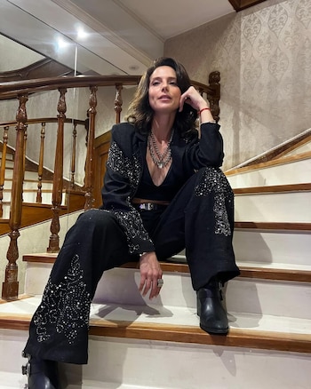 Manuela Pal sentada en una escalera de madera y blanca, vestida con un traje negro con brillo y un top escotado. Tiene el pelo castaño suelto y mira a cámara