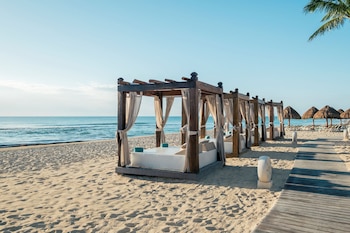 Cabañas de madera con colchones blancos en la arena, un camino de tablas, sombrillas de paja al fondo y el mar abierto bajo el cielo despejado