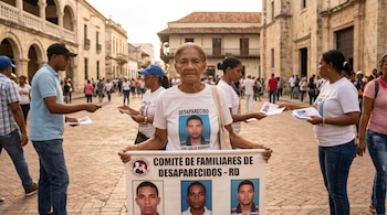 Una anciana con camiseta blanca y otras personas en una calle adoquinada con edificios coloniales, sosteniendo un cartel de desaparecidos y repartiendo folletos.
