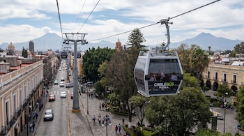 Vista aérea de una cabina de Cablebús en Puebla, México, suspendida sobre una calle con autos y peatones. Al fondo, edificios coloniales y montañas.