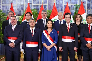 Grupo de hombres y una mujer de pie con banderas de Perú de fondo, algunos usan bandas bicolor rojas y blancas, en una sala formal