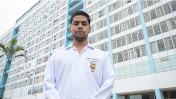 Um jovem de roupas brancas e barba curta, olhando para frente, está em frente a um grande prédio hospitalar branco e azul, com uma palmeira à esquerda.