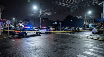Dos patrullas de policía con luces encendidas y cinta de seguridad amarilla acordonan una calle urbana mojada con vehículos estacionados y edificios en la noche.