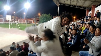 Hombre con camisa blanca es agredido en público, cayendo hacia adelante. Otro hombre lo ataca en primer plano, con espectadores y una cancha deportiva de fondo por la noche.