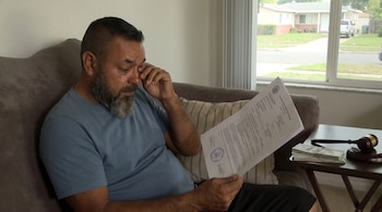 Hombre barbudo con camiseta azul llorando en un sofá, sosteniendo documentos legales, con billetes y un mazo de juez en una mesa a su lado.