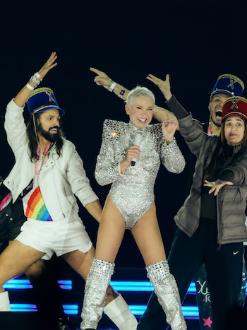 Xuxa con traje plateado, botas altas y micrófono, sonríe en el escenario. La acompañan dos bailarines con sombreros y atuendos vistosos