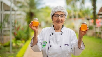 Una mujer mayor con chaqueta de chef, redecilla y anteojos sostiene dos frascos de vidrio con líquido dorado en un jardín