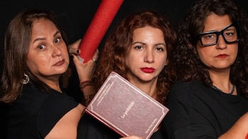Tres mujeres vestidas de negro sostienen un libro de tapa dura titulado Lo prohibido y un objeto de espuma rojo sobre un fondo oscuro