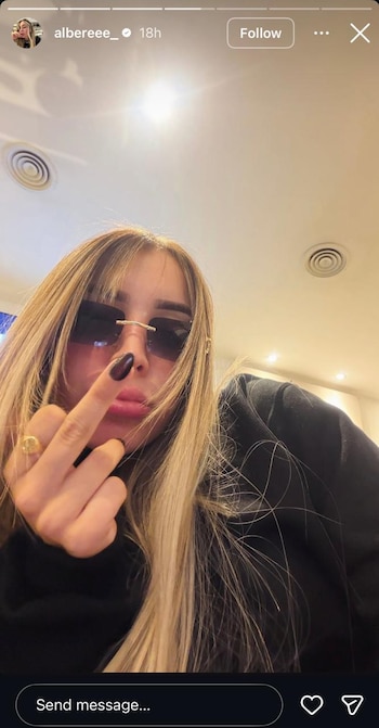 Mujer con cabello rubio, gafas de sol rectangulares oscuras, labial oscuro y uñas negras, levanta el dedo medio. Lleva un anillo dorado. Fondo de techo claro