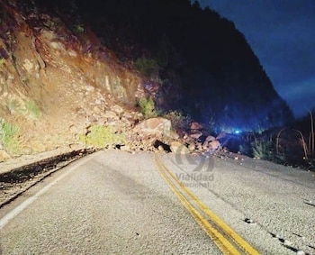 Una carretera asfaltada con doble línea amarilla y rocas grandes en el centro. A la izquierda, una ladera de montaña con vegetación. Noche cerrada