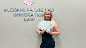 Una mujer rubia con vestido negro sonríe mientras sostiene documentos. Detrás, un letrero con "Alexandra Lozano Immigration Law" y un sello
